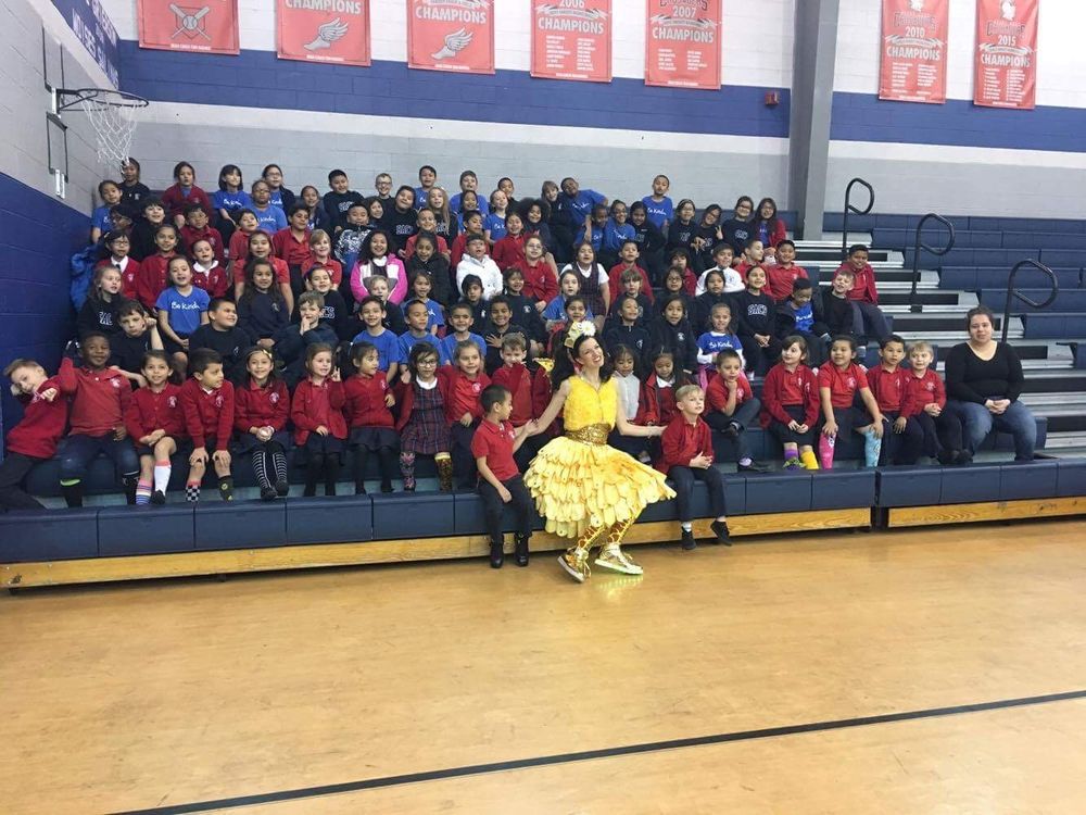 Keri in a yellow fairy costume with a big group of schoolchildren in a gym.