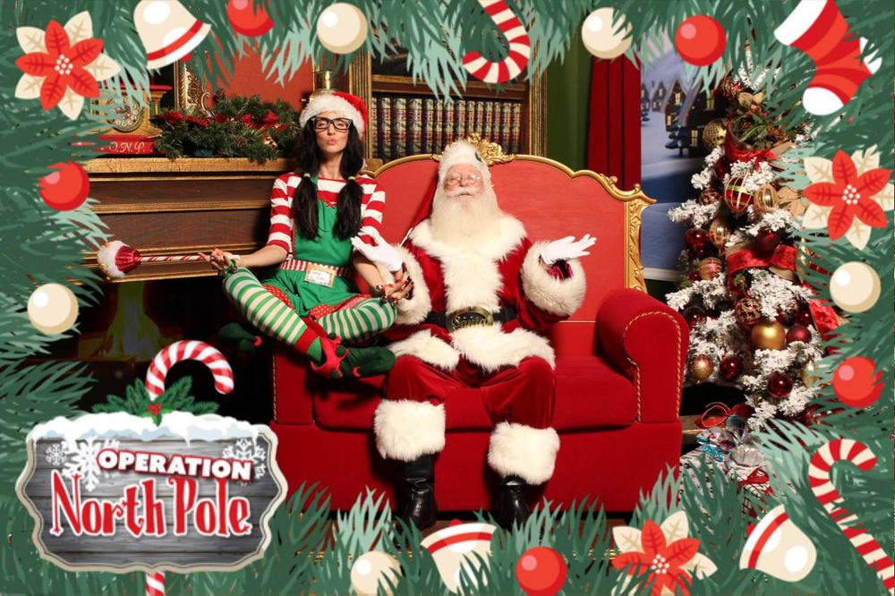 Keri in an elf costume sitting cross-legged beside Santa at a holiday event.