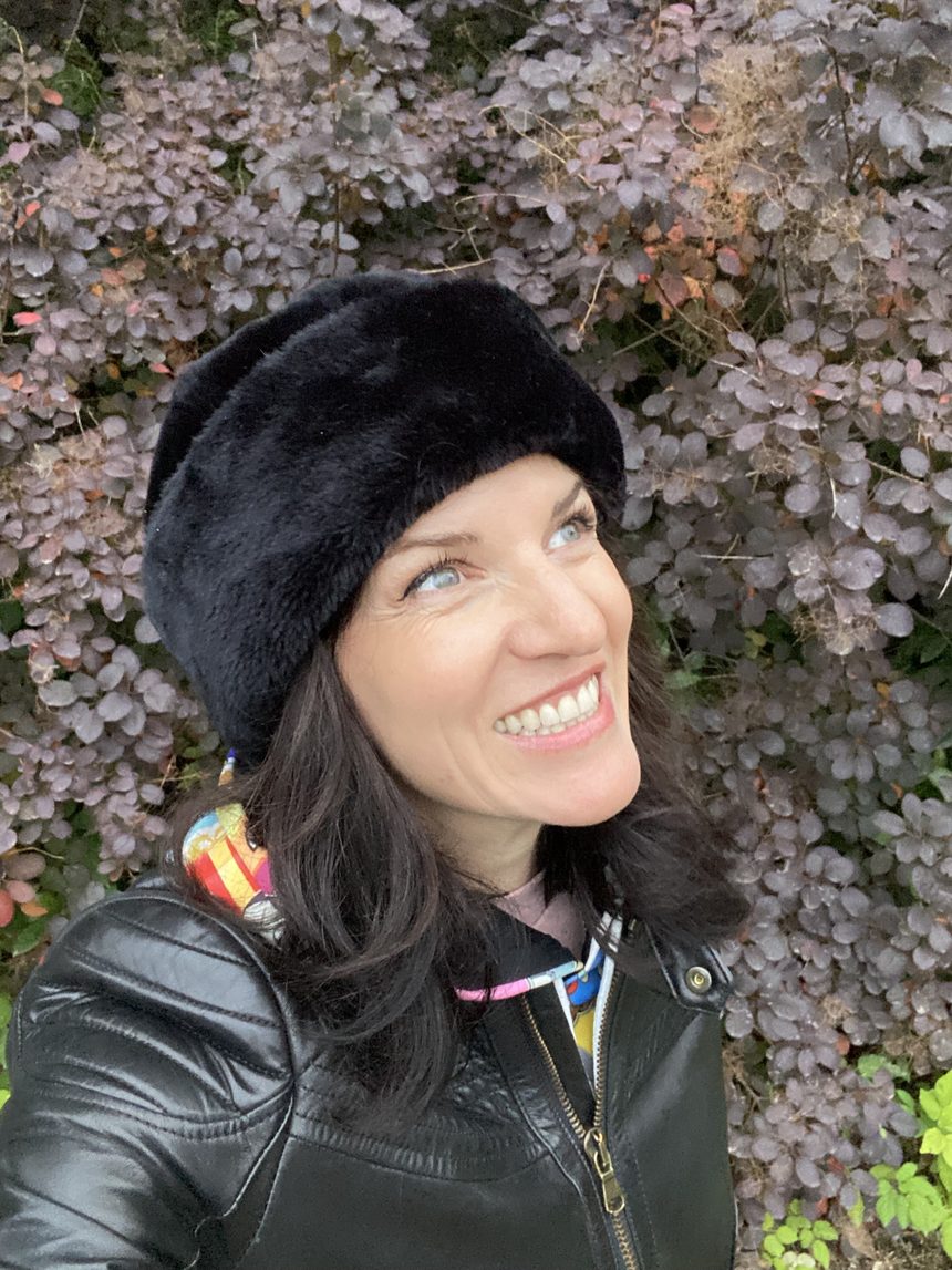Keri smiling brightly in a black beret in front of a wall of purple leaves.